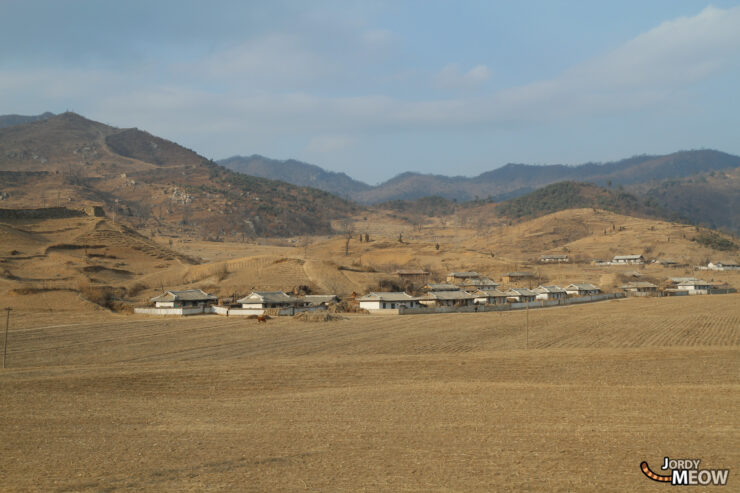 North Korean village surrounded by mountains, fields, and vegetation in a rugged landscape.