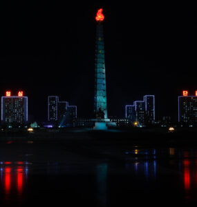 Pyongyang skyline at night with illuminated buildings and tower reflecting in still waters.