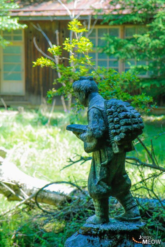 Weathered schoolchild statue reading book in overgrown grass at abandoned Namezawa School near Mt. Fuji