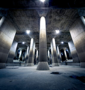 Subterranean flood water diversion channel with towering concrete columns and dramatic lighting.