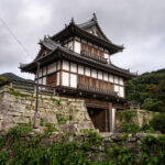 Historic Japanese gate tower on stone wall in Izuhara under cloudy sky
