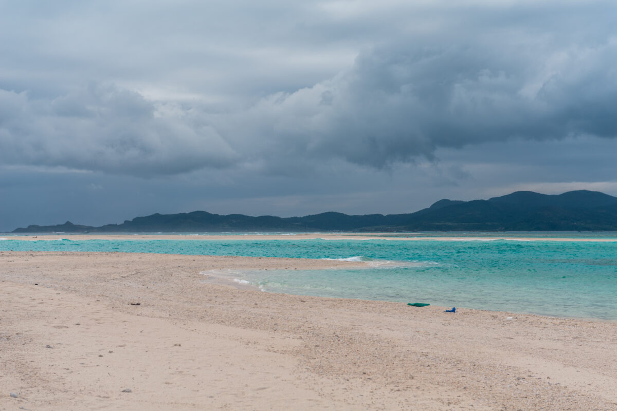 Kumejima beach with pale sand, turquoise shallows, distant ridge, and moody overcast sky