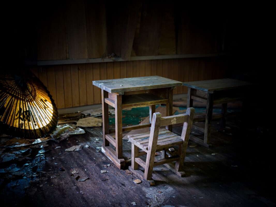 Abandoned Japanese classroom with worn child desk and chair, debris, and dim lantern light.