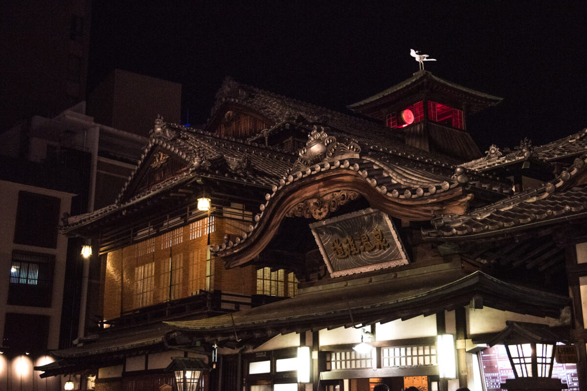 Dogo Onsen bathhouse at night in Matsuyama, Japan, glowing traditional wooden architecture.