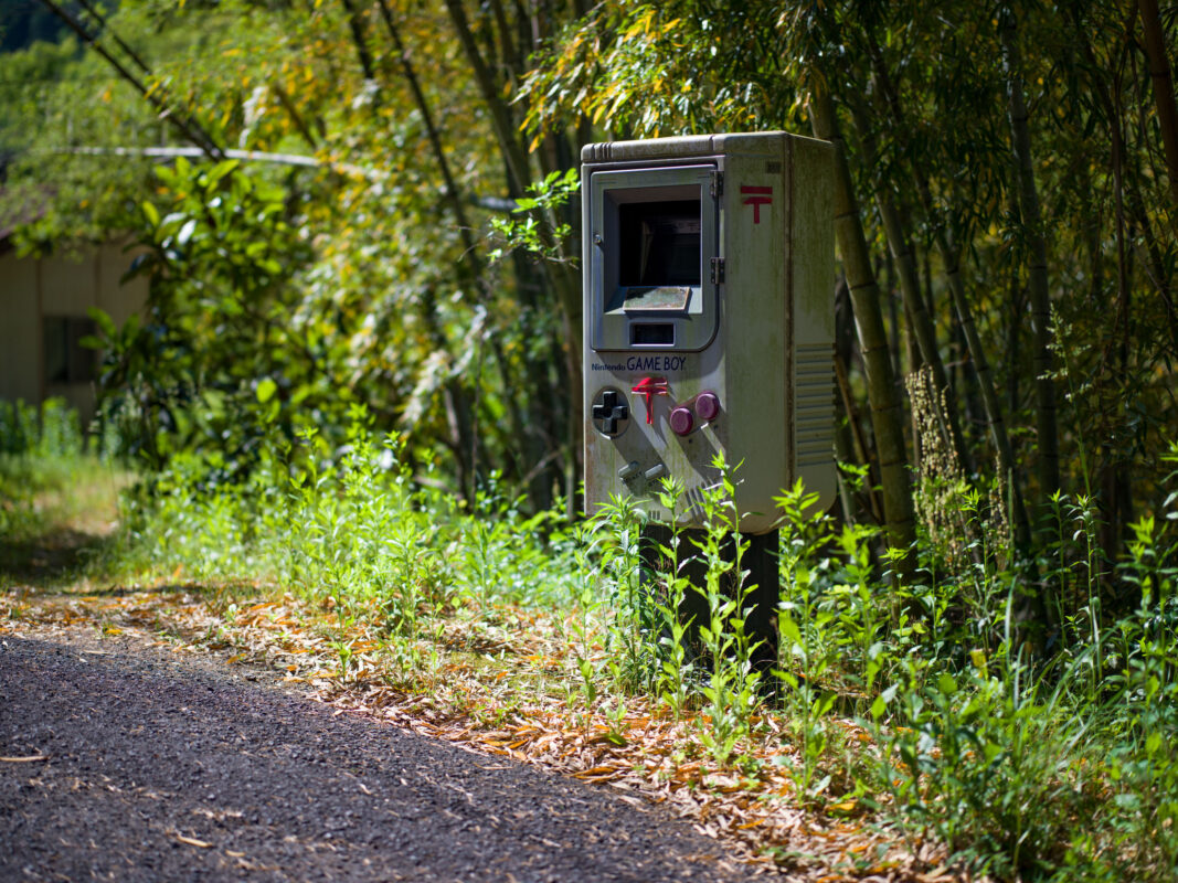 Vintage Game Boy shaped mailbox beside a woodland path with greenery and dappled sunlight